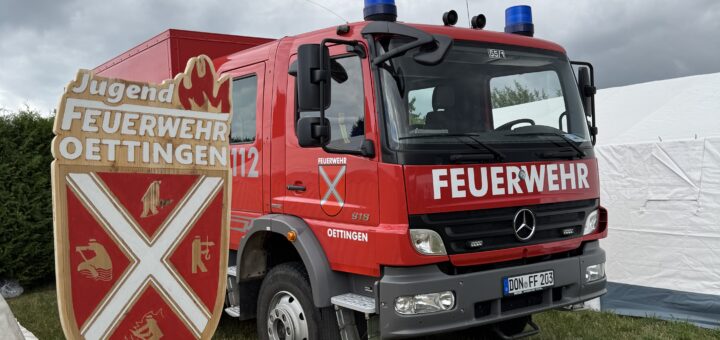 Das Bild zeigt das Schild der Jugendfeuerwehr Oettingen vor einem Oettinger Feuerwehrauto.