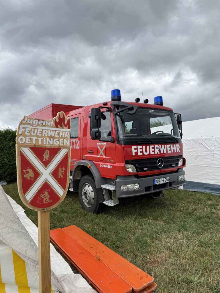 Das Bild zeigt das Schild der Jugendfeuerwehr Oettingen vor einem Oettinger Feuerwehrauto.